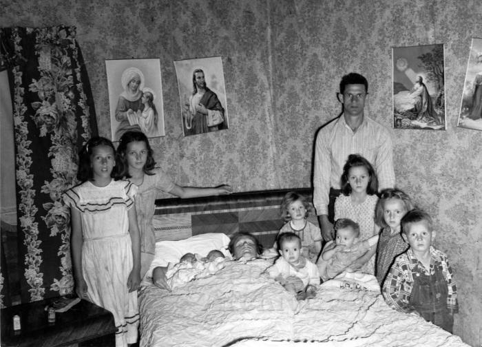 Woman in bed holding 2 infants, surrounded by her husband and children