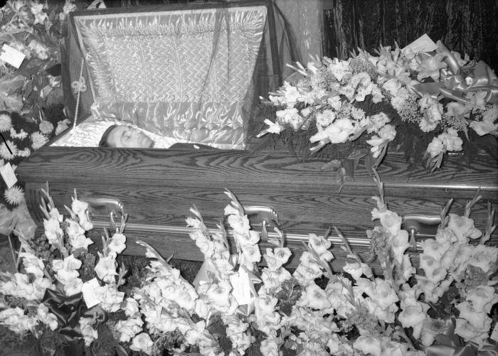 Man in casket surrounded by gladiolus bouquets