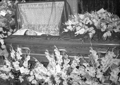 Man in casket surrounded by gladiolus bouquets