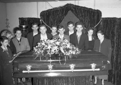 Group portrait in funeral parlor surrounding casket, second version