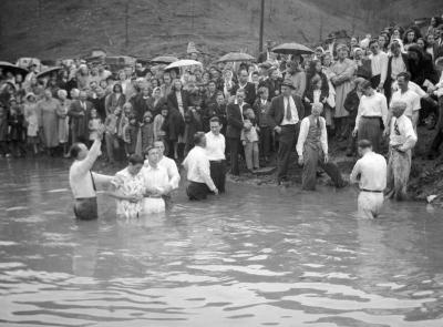 River baptism of two young men, audience holding umbrellas