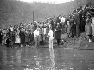 Clergymen performing baptisms stand with crowd along riverbank