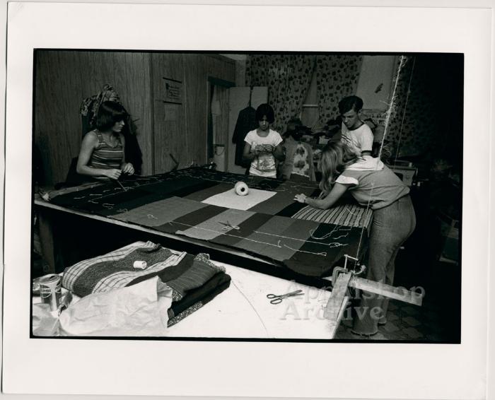 Production still of women and a man working around quilting frame - Millstone Sewing Center