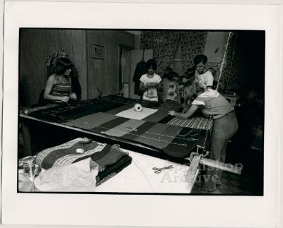 Production still of women and a man working around quilting frame - Millstone Sewing Center